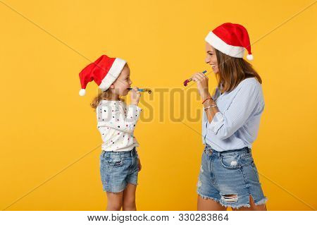 Woman Child Baby Girl In Christmas Santa Hat Blow In Pipe. Mommy Little Kid Daughter Isolated On Yel