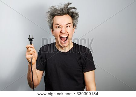 Unshaven Guy With Long Hair Holds An Electric Plug With A Cable In His Hand On A Gray Background