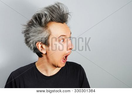 Unshorn And Unshaven Young Guy With Long Hair Surpriced On A Gray Background. Side View.