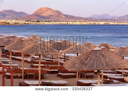 Thatched Peaks Of Beach Umbrellas With Deck Chairs On The Deserted Beach Of The Sea Promenade Agains