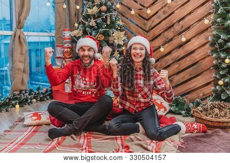 Extremely Happy Man And Woman Are Raising Clunched Fists, Saying Yes, While Sitting Near Christmas T