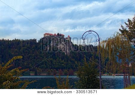 Scenic Landscape Photo Of Bled Castle On The Top Of The Rock And Bled Lake Against Cloudy Sky. Famou