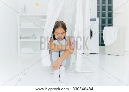 Small Adorable Girl Sits On Floor, Being Punished By Parents, Poses Near Clothes Dryer, Focused Down