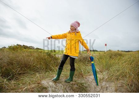 Cheerful Little Girl Staying On Beach With Colorful Ambrella On Baltic Sea At Windy Rainy Weather