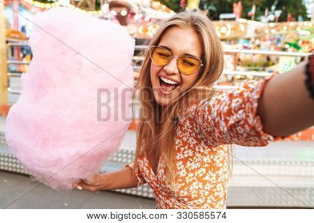 Photo of emotional positive cute woman walking outdoors in amusement park eat candyfloss take selfie by camera.