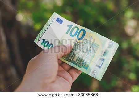 Male Hand Holding 100 Hundredth Euro Banknote On Green Background Outdoors, Closeup