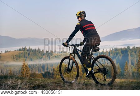 Male Cyclist In Cycling Suit Riding Bike With Coniferous Trees And Hills On Background. Man Bicyclis
