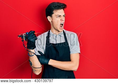 Young hispanic man tattoo artist wearing professional uniform and gloves holding tattooer machine pointing with finger to the camera and to you, confident gesture looking serious 