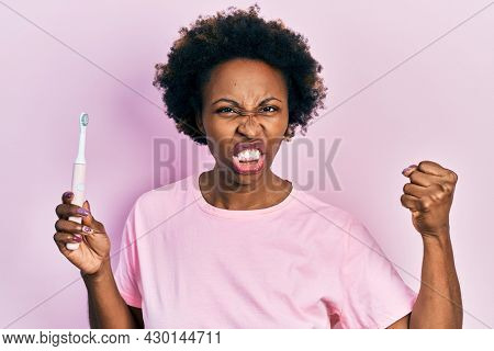 Young african american woman holding electric toothbrush annoyed and frustrated shouting with anger, yelling crazy with anger and hand raised 