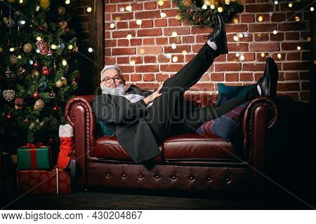 Funny mature man with gray beard and gray hair posing making faces lying on the sofa at home decorated for Christmas. Merry Christmas and Happy New Year! 