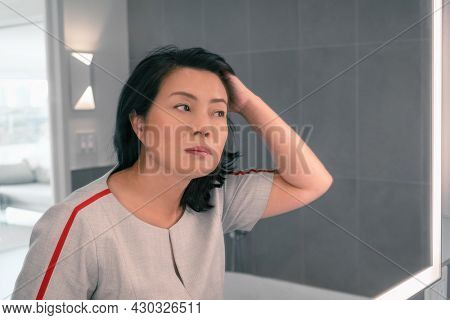 Hair loss mature Asian woman touching her hair styling or coloring gray hair looking at herself in home mirror. Beautifiul chinese professional lady makeup.