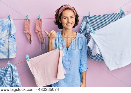 Young brunette woman doing laundry around string hangs smiling with happy face looking and pointing to the side with thumb up. 