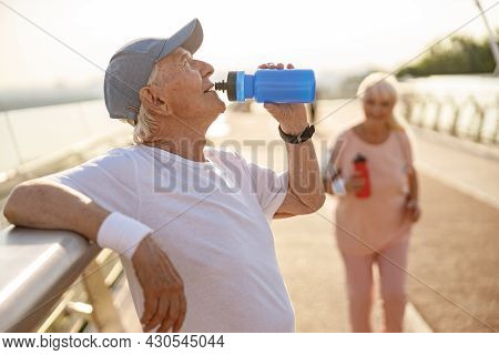 Positive Senior Man Drinks Water Leaning Onto Railing While Lady Runs Along Footbridge
