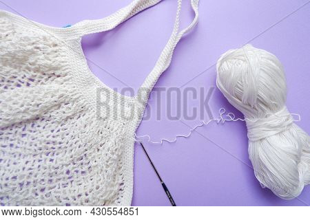 Eco Bag In The Knitting Process. White Grid Bag, Crochet Hook And Remaining Yarn On A Purple Backgro
