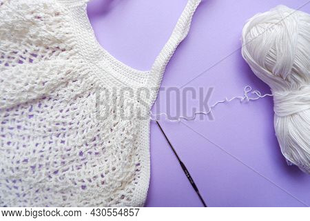 Eco Bag In The Knitting Process. White Grid Bag, Crochet Hook And Remaining Yarn On A Purple Backgro