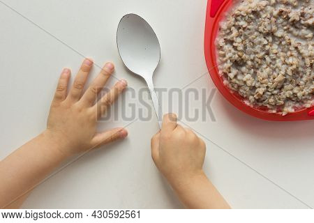 Top View Of Child Hands Holding Spoon In Right Hand And Buckwheat Porridge On White Background