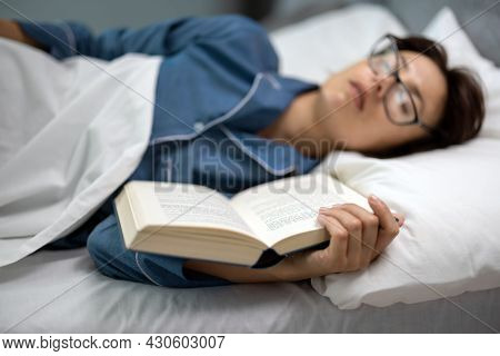 Middle Aged Woman In Eyeglasses And Pajams Enjoying Sweet Nap On Comfy Bed With Book In Hands. Cauca