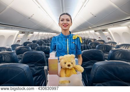 Attractive Female Air Hostess In Bright Blue Uniform Smiling At Camera While Leading Trolley Cart Wi