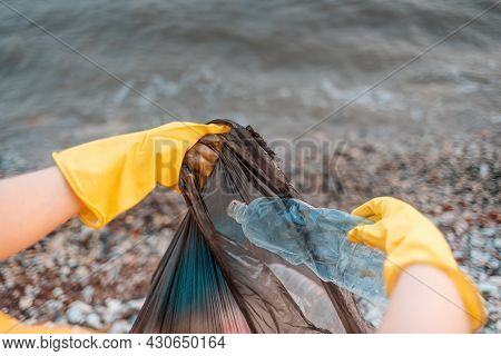 Close Up Of Volunteer's Hands In Rubber Gloves Puts A Dirty Plastic Bottle In Bag With Garbage. Ocea
