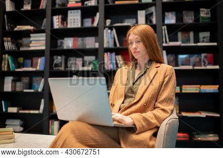 Young female with laptop looking through e-book on screen while sitting in bookshop