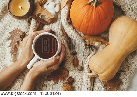 Hands Holding Warm Cup Of Tea On Stylish Cozy Sweater With Pumpkins, Autumn Leaves, Burning Candle. 