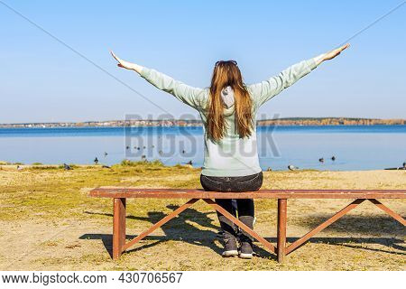 Hipster Young Woman Sitting On Bench By Lake Shore And Pulls Hands To Sun, Zen Like On Autumn Sunny 