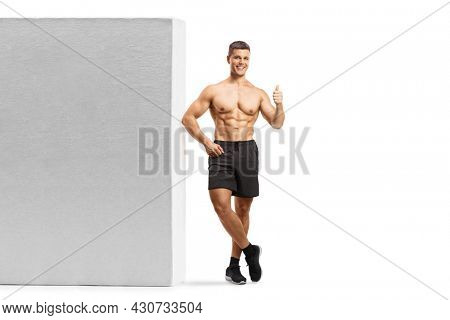 Full length portrait of a bodybuilder leaning shirtless on a wall and showing a thumb up sign isolated on white background