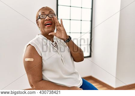 Mature hispanic woman getting vaccine showing arm with band aid shouting and screaming loud to side with hand on mouth. communication concept. 