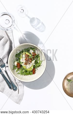 Avocado and stracciatella cheese salad. Fancy dinning with Italian salad bowl on white table with simple contemporary decor. Sunlight and harsh shadow still life. Avocado, cheese and tomato salad