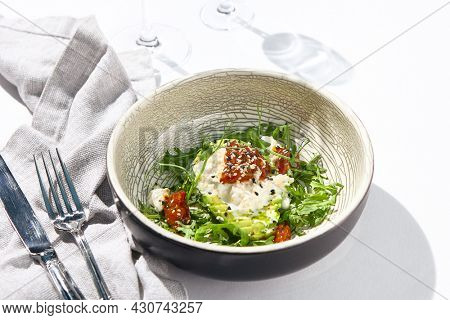 Avocado and stracciatella cheese salad. Fancy dinning with Italian salad bowl on white table with simple contemporary decor. Sunlight and harsh shadow still life. Avocado, cheese and tomato salad