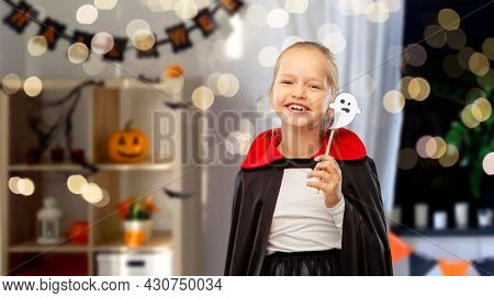 halloween party, holiday and childhood concept - happy smiling girl in black dracula costume with cape and ghost over lights and decorated home room background