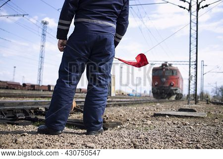Railroader Or Switchman At Station Waving With Red Flag To The Incoming Train And Signalizing Locomo