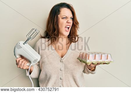 Young caucasian woman holding pastry blender electric mixer and eggs angry and mad screaming frustrated and furious, shouting with anger. rage and aggressive concept. 