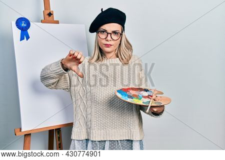 Beautiful caucasian woman drawing with palette on easel stand winner first place with angry face, negative sign showing dislike with thumbs down, rejection concept 