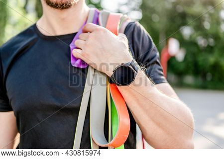 Sportive man guy holding in his hand wearing fitness tracker set of colorful elastic rubber bands at the stadium outdoors. Athlete male person during workout with additional sport equipmant
