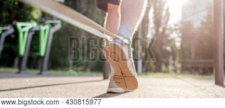 Closeup back view of legs feets of man guy training with elastic rubber band at the stadium outdoors. Adult male person during workout with additional sport equipmant with sunlight
