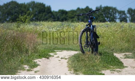 Bike Stands On The Road In The Field. A Mountain Bike Stands On A Field Path With Green Grass. Mount