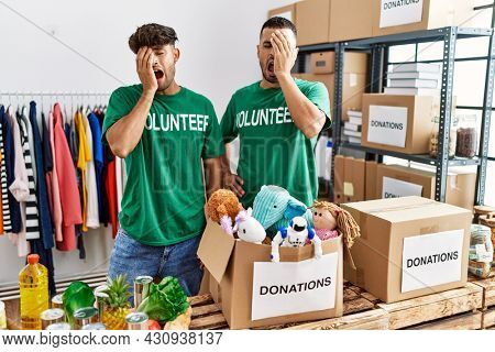 Young gay couple wearing volunteer t shirt at donations stand yawning tired covering half face, eye and mouth with hand. face hurts in pain. 