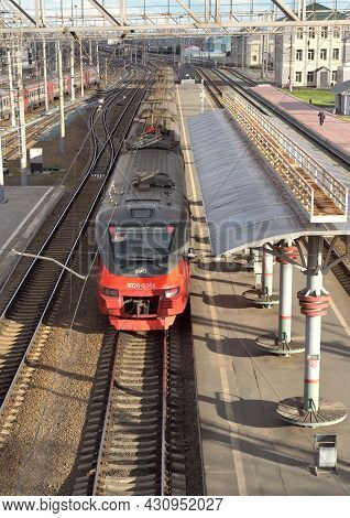 The Modern Red Train Stands On The Tracks At The Passenger Covered Empty Platform, The Railway Track