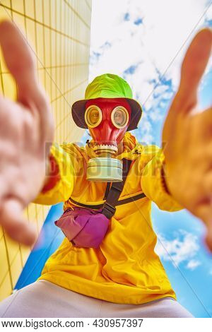 Man in bright colored clothes and a red gas mask looks at camera against a blue sky and yellow industrial wall. Technological disaster concept, air pollution. 