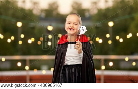 halloween, holiday and childhood concept - happy smiling girl in black dracula costume with cape and ghost over garland lights at roof top party background