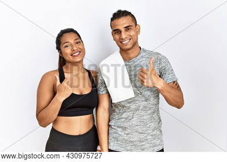 Young latin couple wearing sportswear standing over isolated background doing happy thumbs up gesture with hand. approving expression looking at the camera showing success. 