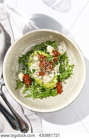 Avocado and stracciatella cheese salad. Fancy dinning with Italian salad bowl on white table with simple contemporary decor. Sunlight and harsh shadow still life. Avocado, cheese and tomato salad