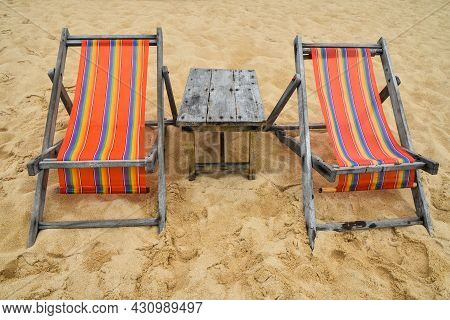 Two Empty Colorful Canvas Lounge Sling Chairs And Wooden Table On Sand Beach Of Resort In Thailand, 