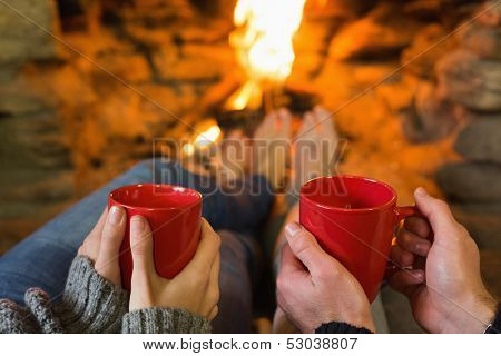 Close-up of hands holding red coffee cups in front of lit fireplace