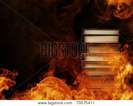 Conceptual image of a tall stack of hardcover books in a burning fire with flames and smoke swirling around them in a darkened room with copyspace