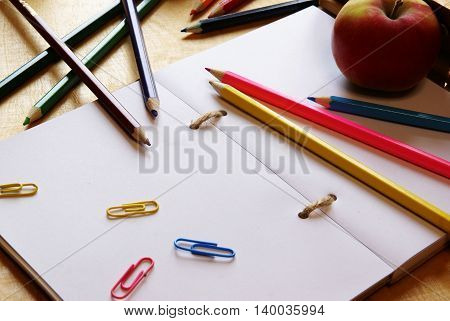 Back to school. Notepad with white sheets, color pencils, staples and fresh red apple on a wooden table. Bright colors.