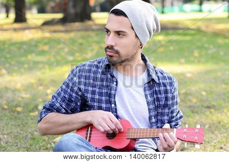 Portrait of young handsome man playing the ukelele in a park. Outdoors.