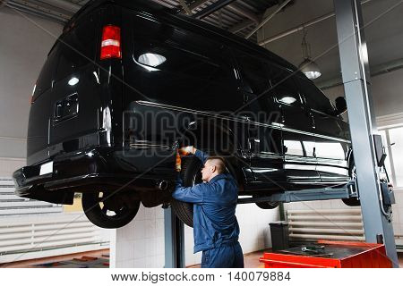 Black minibus on repair in garage. Mechanic working with SUV, lifted on elevator at modern auto service station
