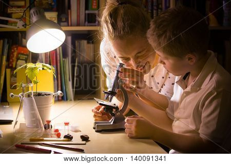 Young mother and elementary school kid boy looking into microscope at home. Family studying samples under the microscope. Science activities with children. Preparing for science lesson.